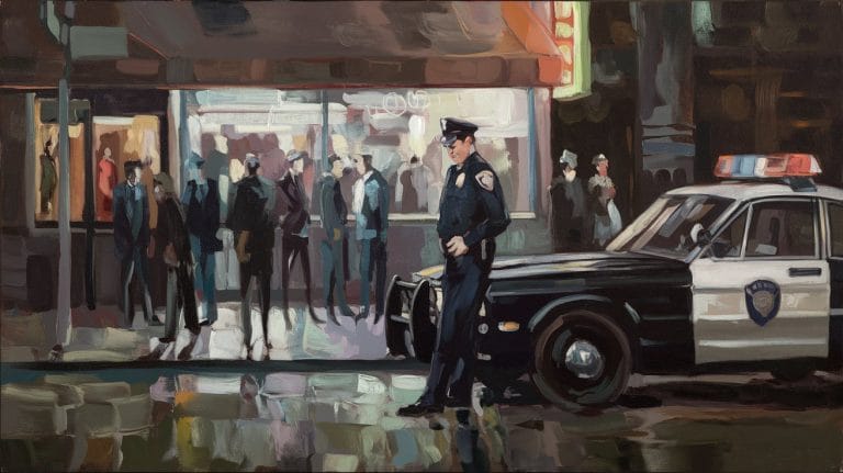 A policeman stands next to a patrol car in front of a busy, brightly lit storefront at night while a crowd looks on.